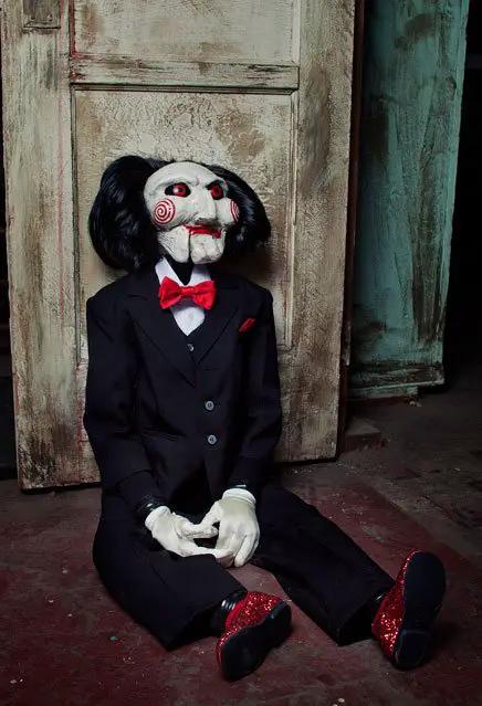 Saw Billy 119 cm doll from TRICK OR TREAT STUDIOS in a black tuxedo with red bow tie, seated on a worn floor.